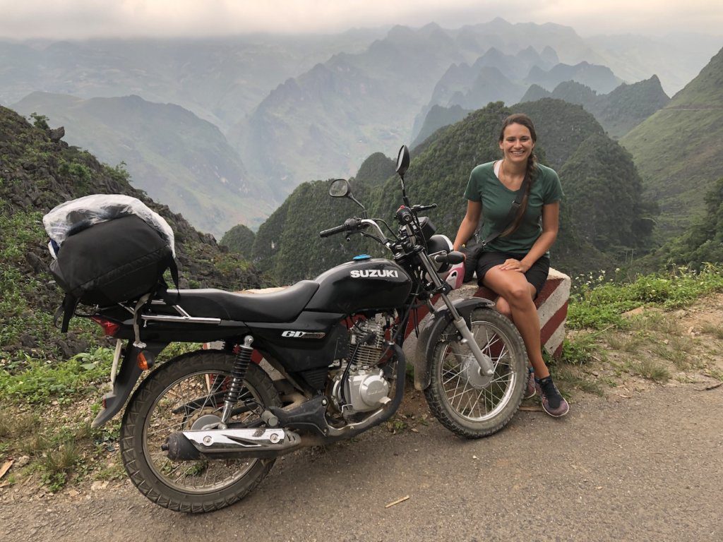 Nicola sits next to a motorbike on the Ha Giang Loop with mountains in the background. She is smiling and wearing a green tee shirt