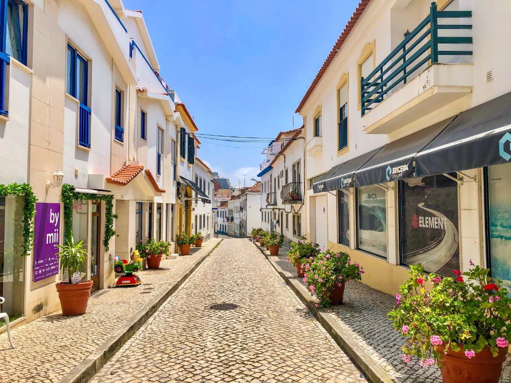 A sunny cobblestone street with flowers along the sidewalk in Ericeira