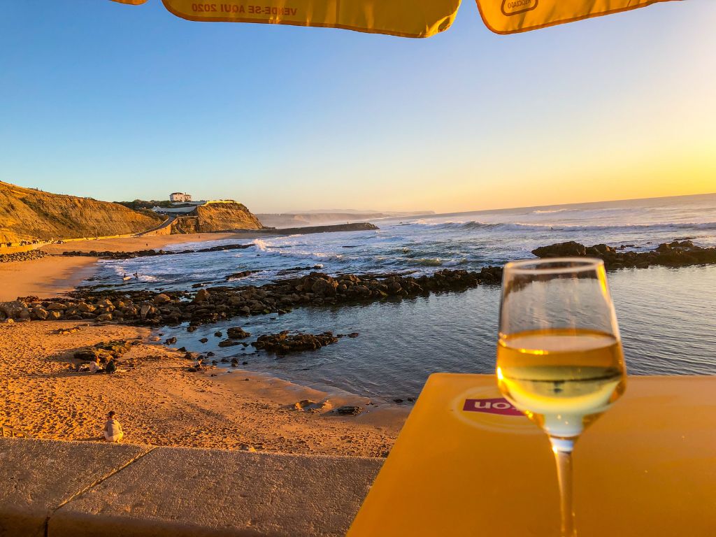 A glass of white wine on a yellow table with Praia do Sul in the background, close to sunset