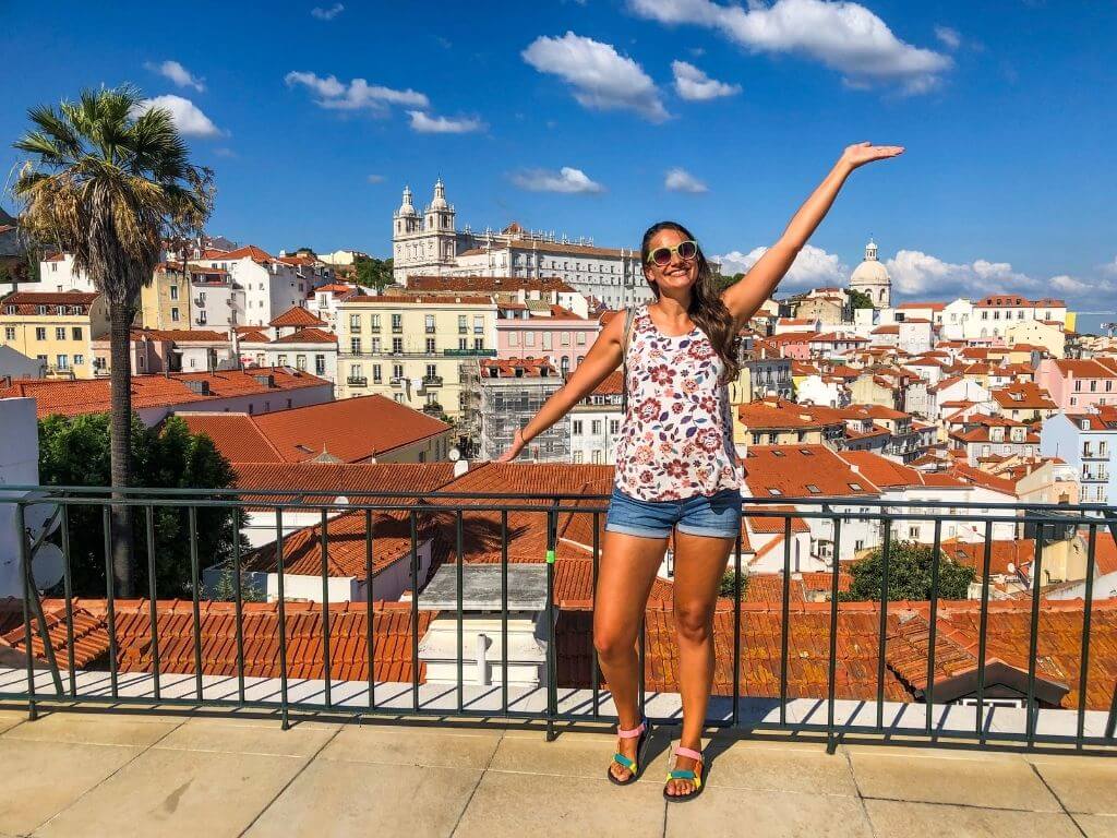 Nicola stands at a lookout point with her arms outstretched. Behind her is a city view of Lisbon and a palm tree