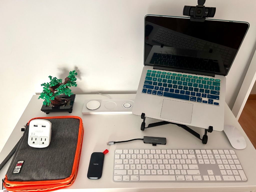 A digital nomad office setup with a travel electronics bag, laptop, laptop stand, wireless keyboard, wireless mouse, webcam, charging station, and external hard drive on a white table top