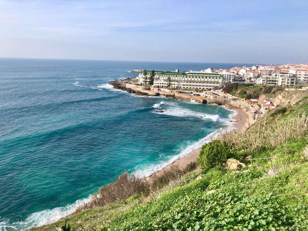 The blue water of Ericeira below a cliff with grass and plants on it, The Ericeira World Surf Reserve is a hidden gem in Portugal